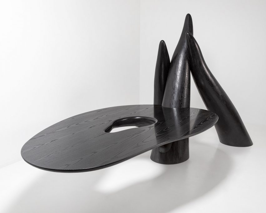 Foto objek patung berwarna hitam dengan permukaan seperti meja besar yang memanjang dari tiga pilar mirip tentakel yang menjulang dari lantai.